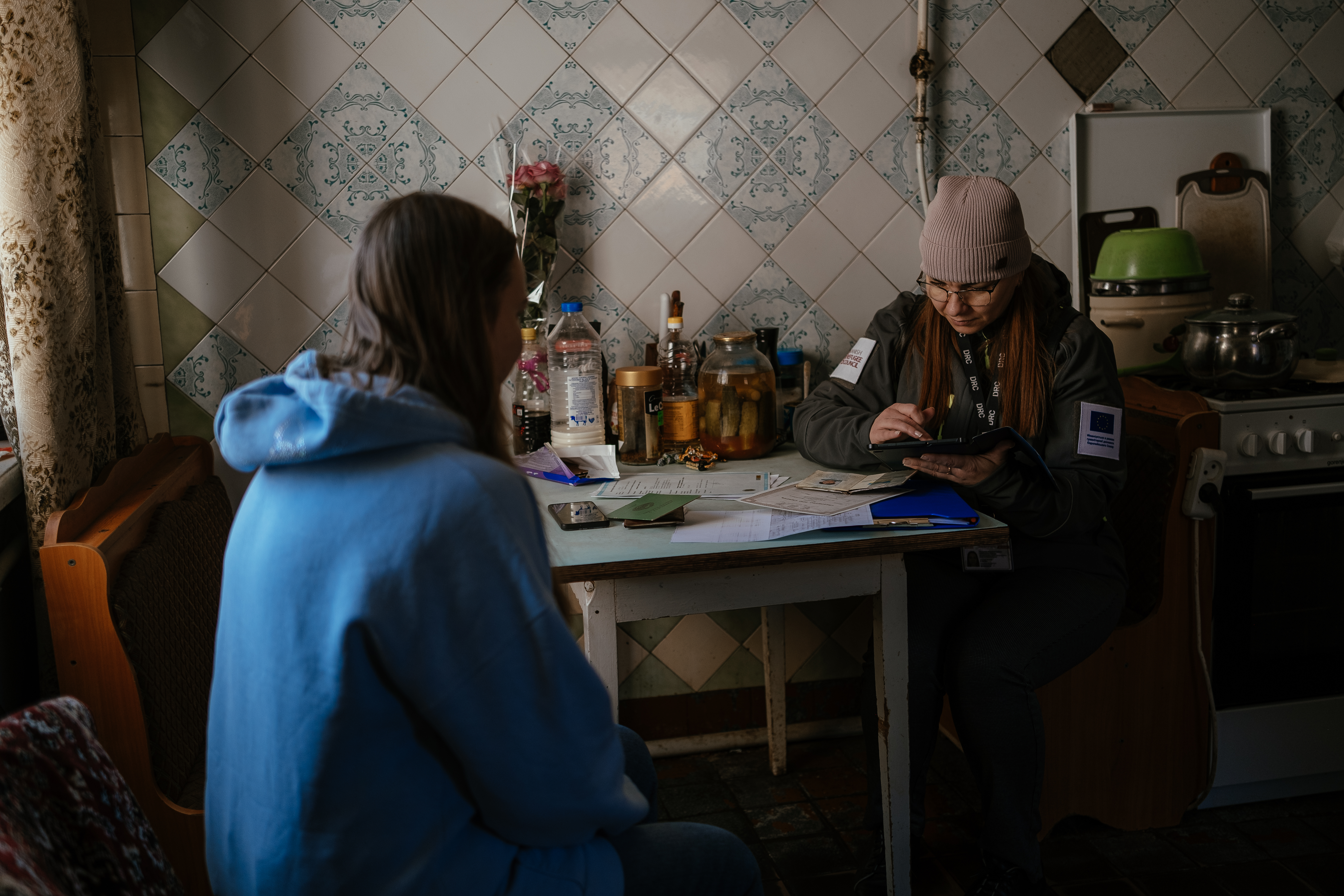 Liudmyla, DRC Shelter and Settlement assistant, sits in Svitlana’s kitchen, registering the woman for humanitarian assistance. ©DRC Ukraine, Cherkaska Lozova, Kharkiv Oblast, March 2025, Krystyna Pashkina