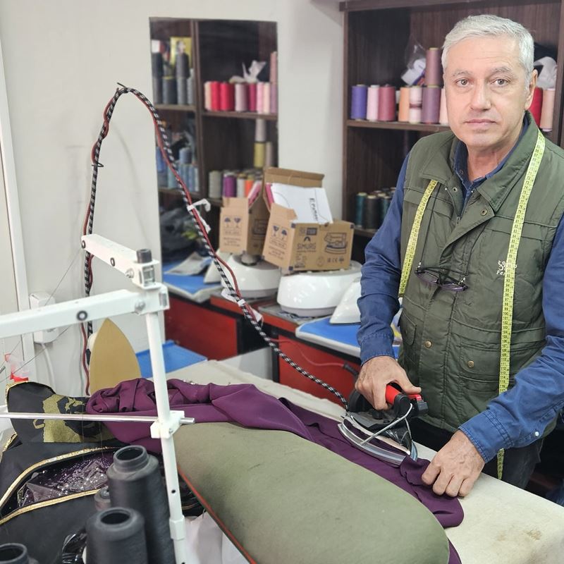 Mehmet is standing at his workbench inside his small shop. He is ironing a deep purple garment.